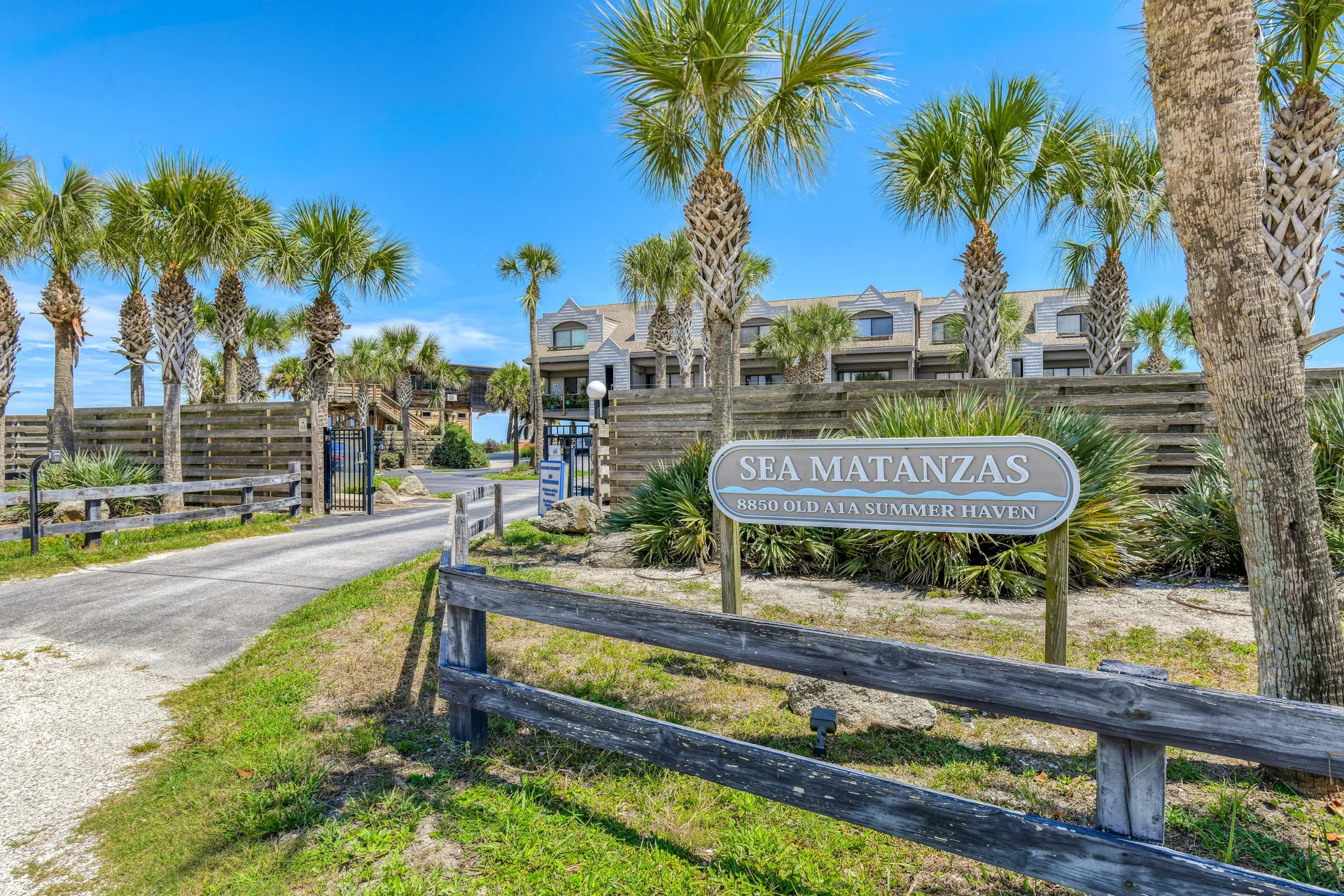 Entrance to beachfront condos marked by a wooden sign, palm trees, and a gated driveway under a bright blue sky.