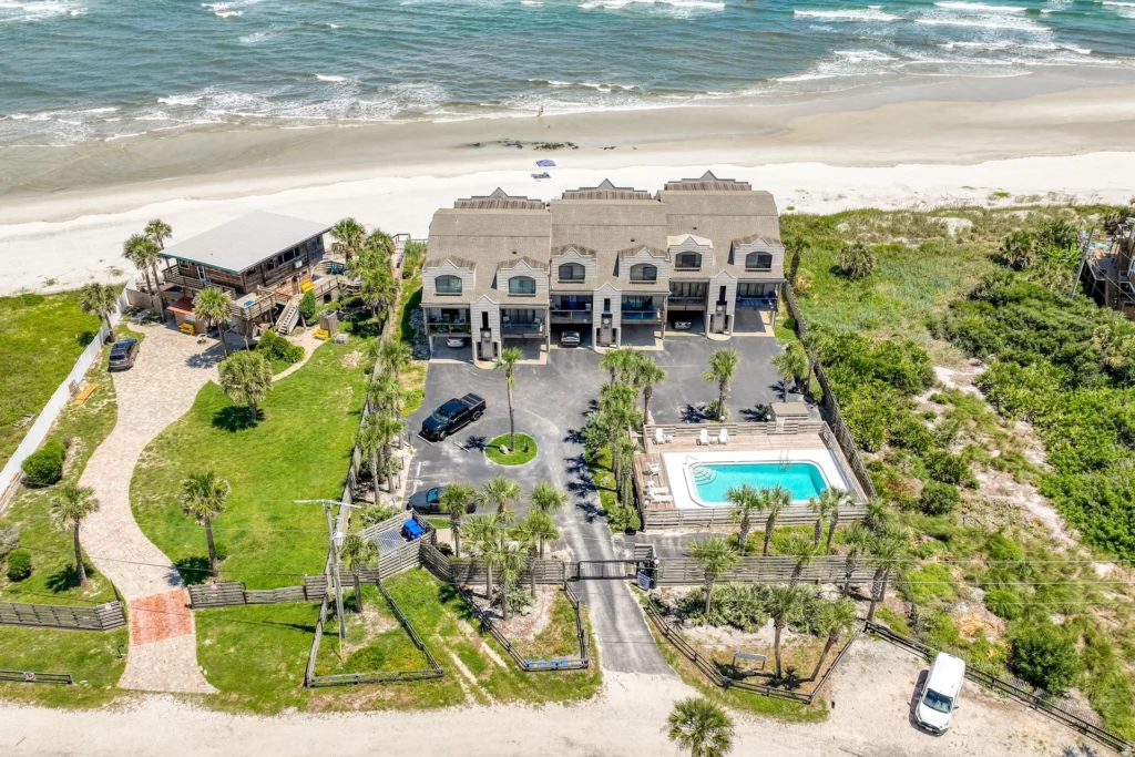 Gated beachfront condos with asphalt parking, fenced communal pool, and green lawns leading to sandy shore.