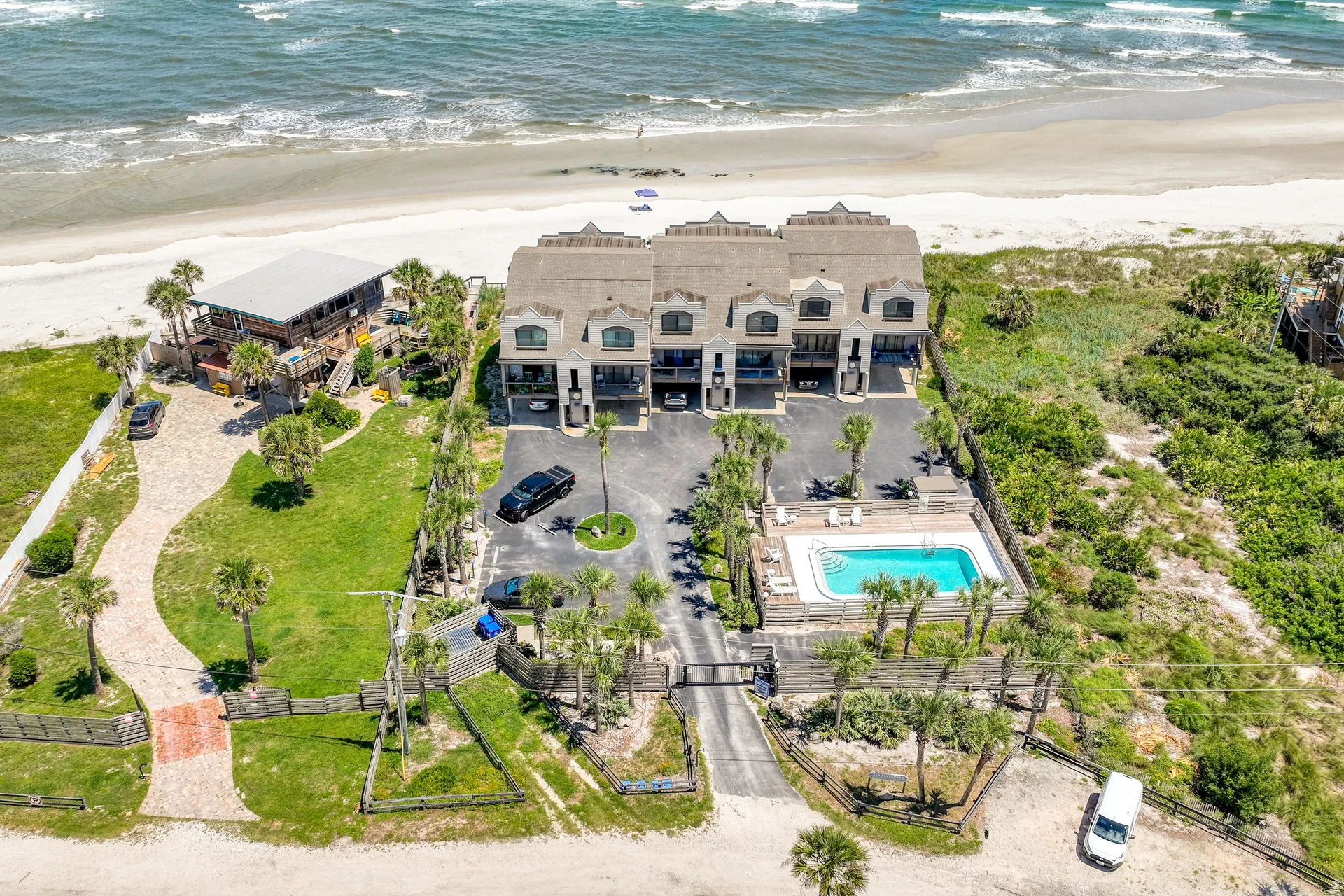 Gated beachfront condos with asphalt parking, fenced communal pool, and green lawns leading to sandy shore.