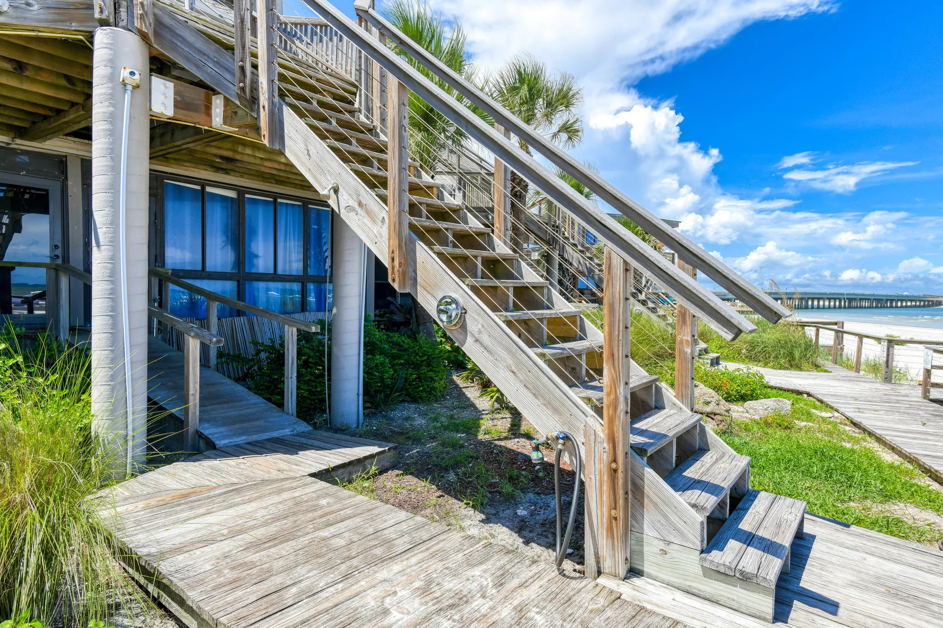 Exterior wooden stairs with cable railing lead down to a sandy beach and boardwalk under a partly cloudy sky.