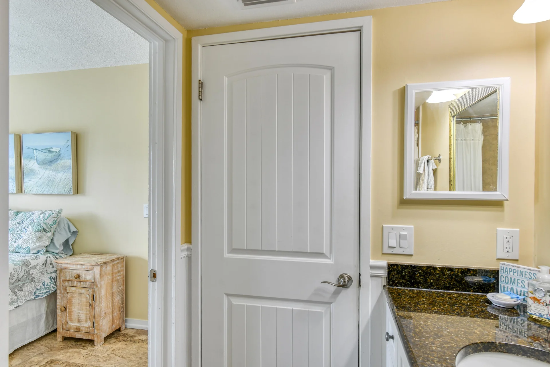 Bathroom vanity with dark granite countertop, white framed mirror, and a glimpse into the adjacent bedroom with coastal artwork.