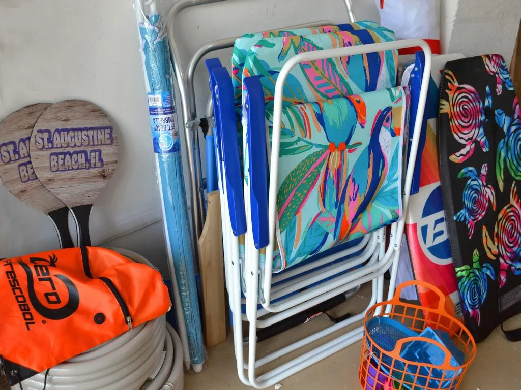 Beach gear storage with colorful folding chairs, wooden paddleball rackets, an orange basket, and a coiled hose.