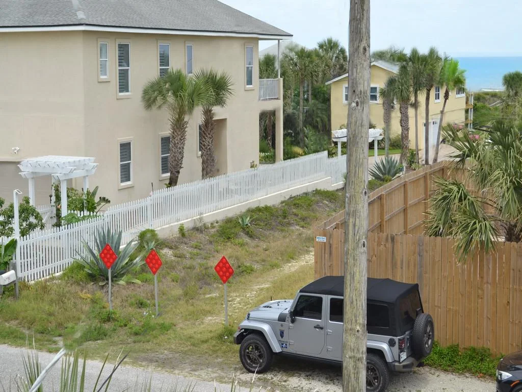 View from balcony overlooking sandy path, palm trees, neighboring houses, and a glimpse of the ocean horizon.