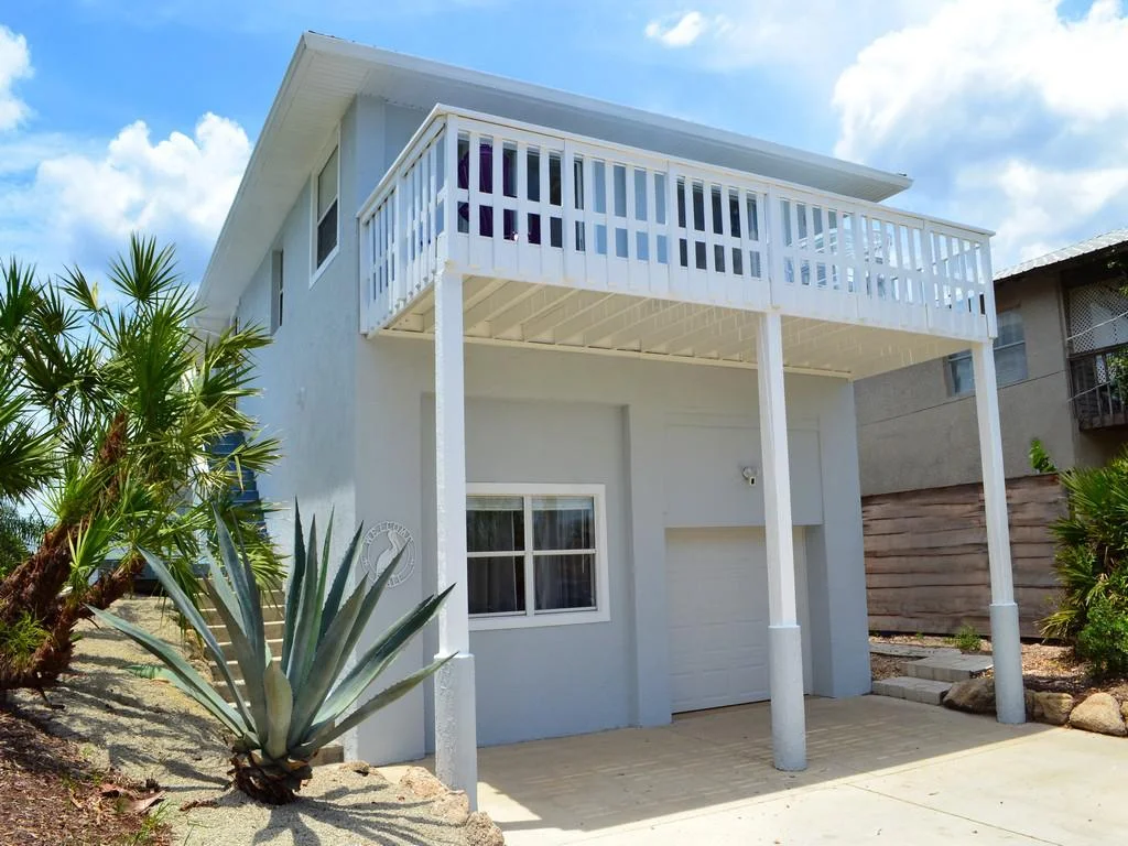 Two-story gray house with white balcony railing, concrete driveway, and desert landscaping featuring agave and palm plants.