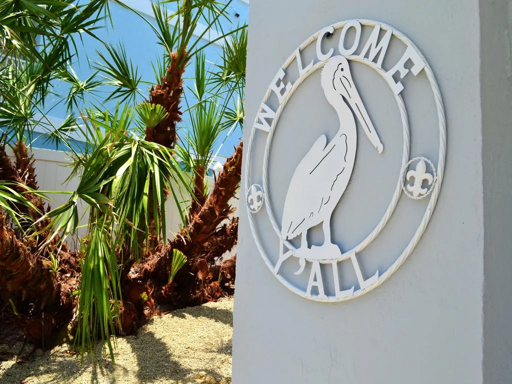 White metal pelican wall art with "Welcome Y'all" text mounted on stucco near tropical palm plants.