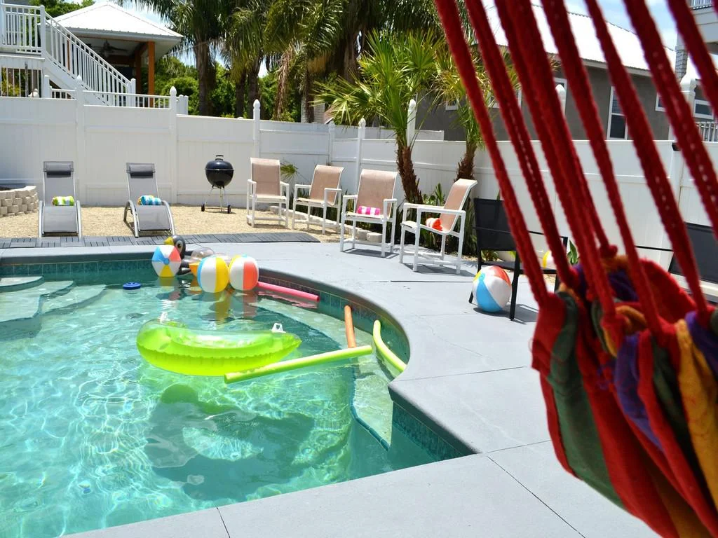 Colorful hammock ropes frame the gray concrete pool deck with beige mesh lounge chairs and beach balls floating in the water.