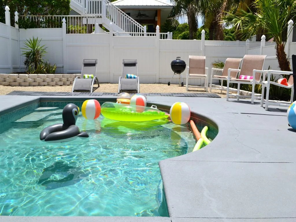 Concrete pool deck with inflatable toys floating in clear water, surrounded by beige mesh chairs and a black charcoal grill.