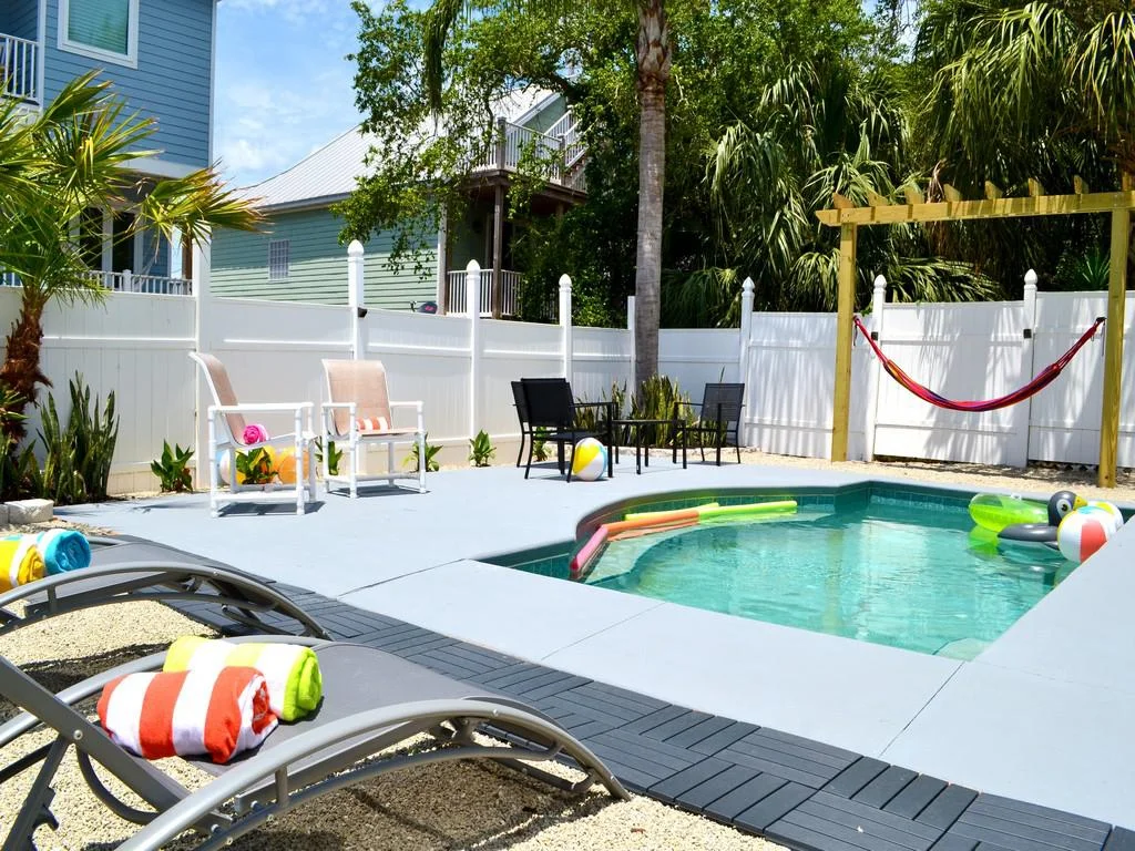 Small pool with gray concrete deck, surrounded by white fencing, loungers with striped towels, and a red hammock.