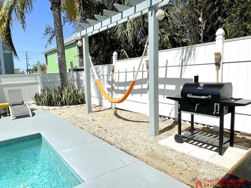 Concrete pool deck surrounds turquoise pool water, with a white pergola holding an orange hammock and a black charcoal grill nearby.