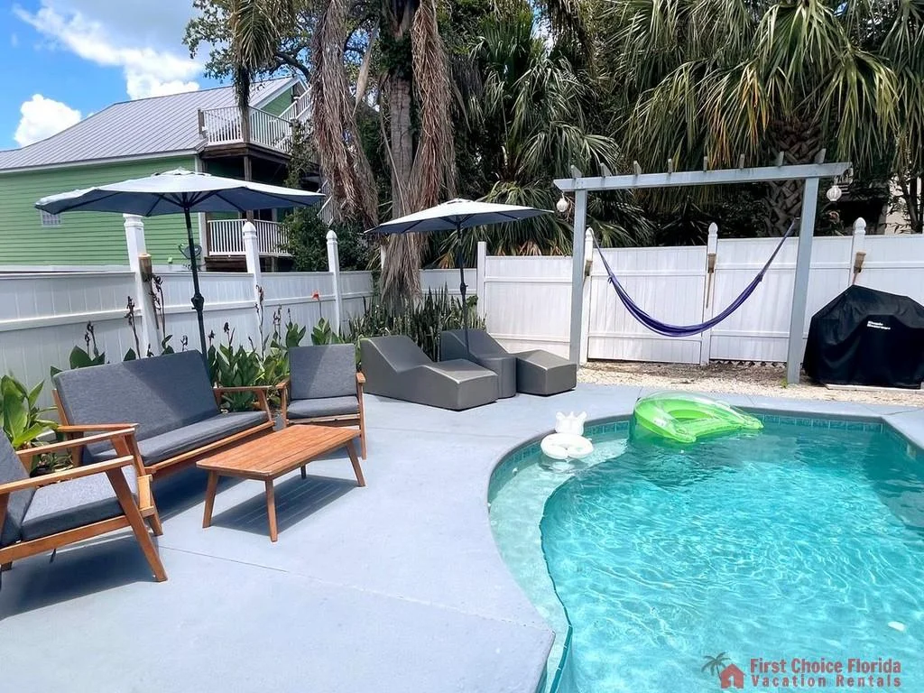 Poolside seating includes gray cushioned wood chairs, a wooden coffee table, and two gray lounge chairs under umbrellas.