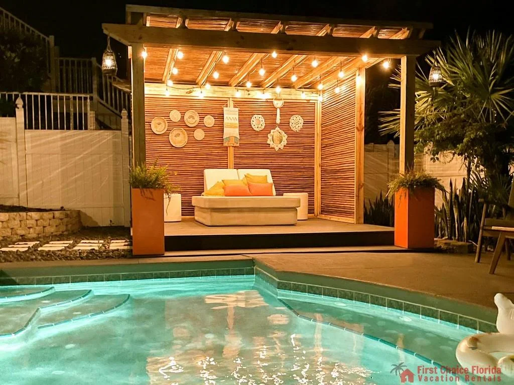 Nighttime view of illuminated pool with curved steps and a wooden pergola featuring a white cushioned loveseat and woven wall decor.