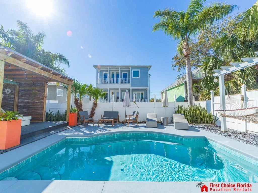 Curved-edge pool with light blue tile border, surrounded by gray deck, lounge chairs, and a hammock under a white pergola.