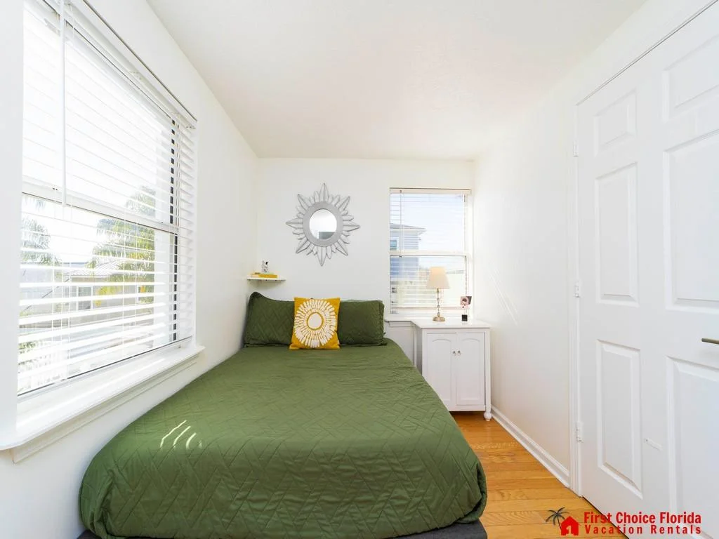Natural light fills the bedroom with wood floors, a white corner cabinet, and a decorative silver sunburst mirror above the bed.