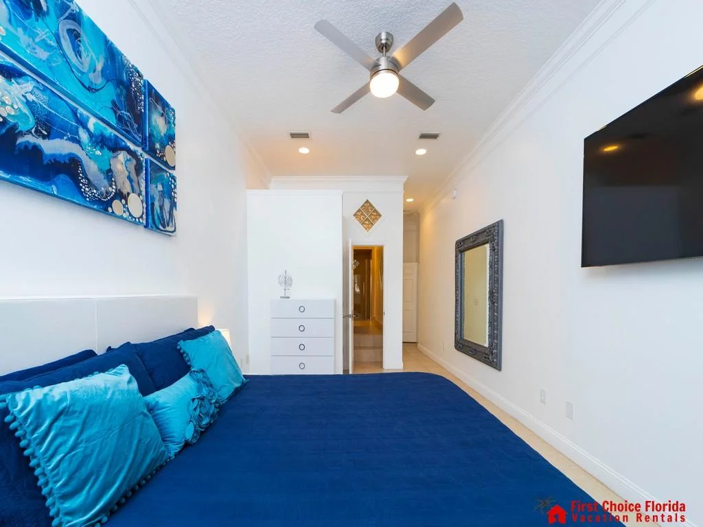 Bedroom features a navy bedspread with blue accent pillows, a white dresser, wall-mounted TV, and a ceiling fan.