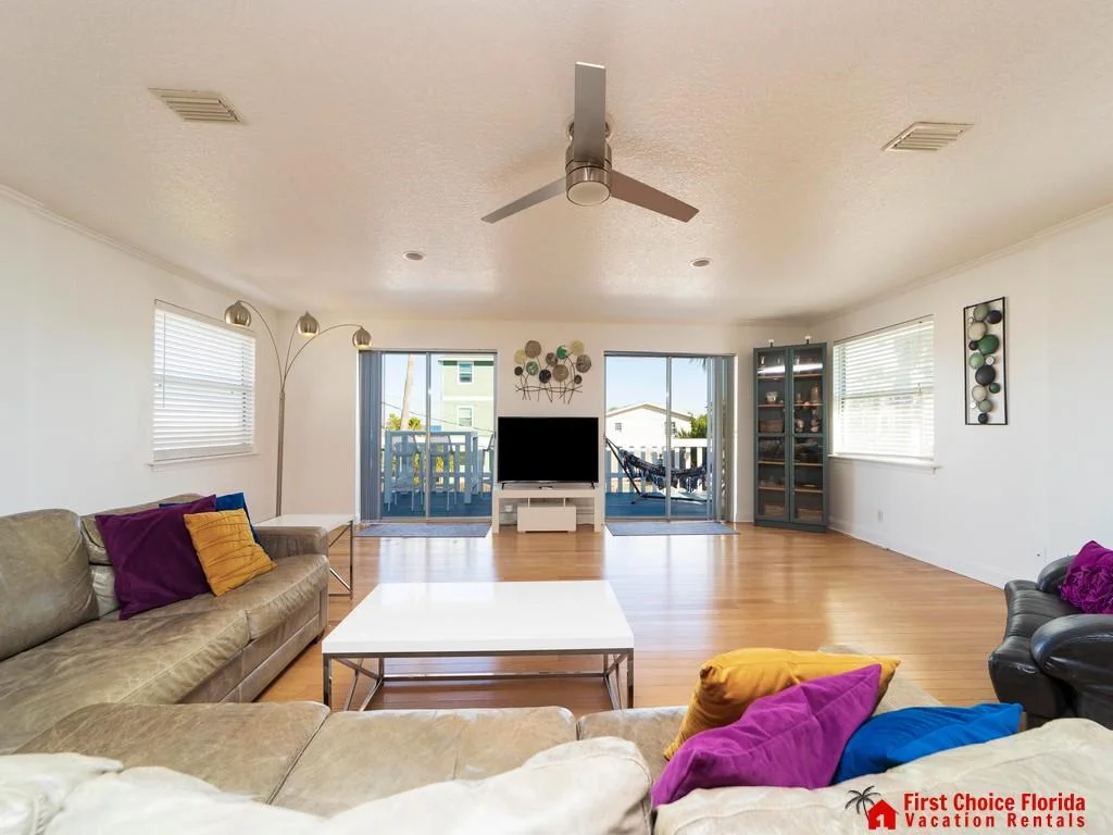 Living room with light wood floors, leather sofas accented by colorful pillows, and sliding doors opening to a balcony.