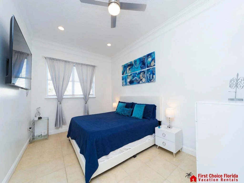 Bedroom with white leather bed frame, navy bedding, blue abstract wall art, sheer curtains, and mounted flat-screen TV.