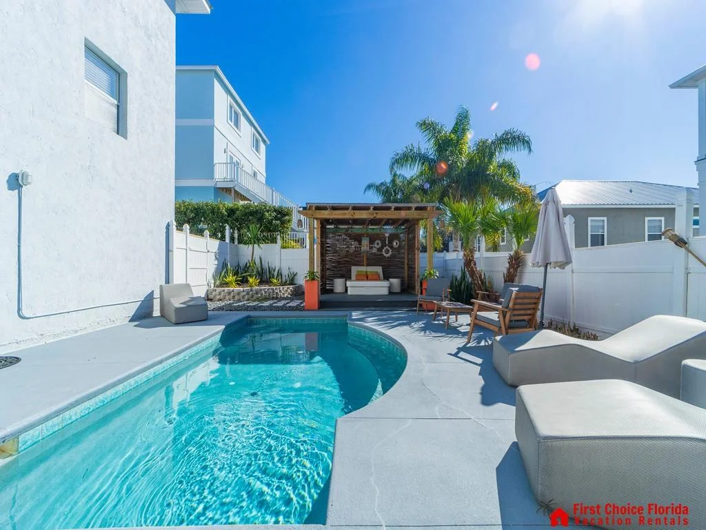 Curved-edge pool with clear water, surrounded by gray concrete deck, wood pergola, and cushioned seating.