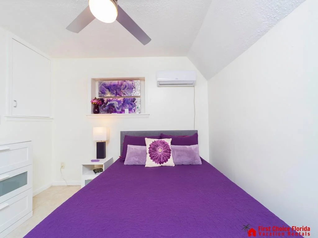 Purple bedspread with decorative pillows centered under a wall-mounted air conditioner and a small window with purple artwork.