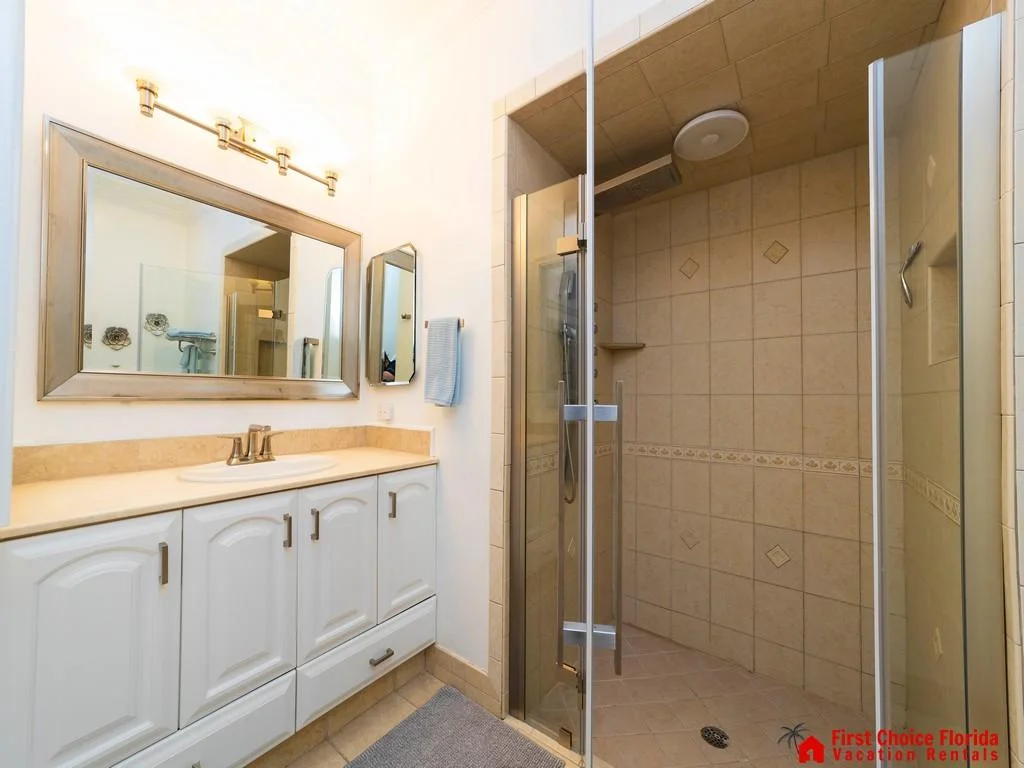 Bathroom with beige tiled walk-in shower, glass door, white vanity, and large framed mirror under brass lighting.
