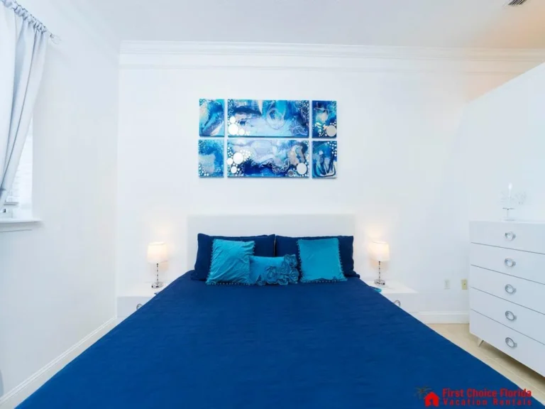 Bedroom features a navy quilted bedspread, turquoise accent pillows, white nightstands with lamps, and abstract blue wall art.