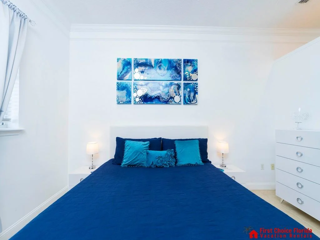 Bedroom features a navy quilted bedspread, turquoise accent pillows, white nightstands with lamps, and abstract blue wall art.