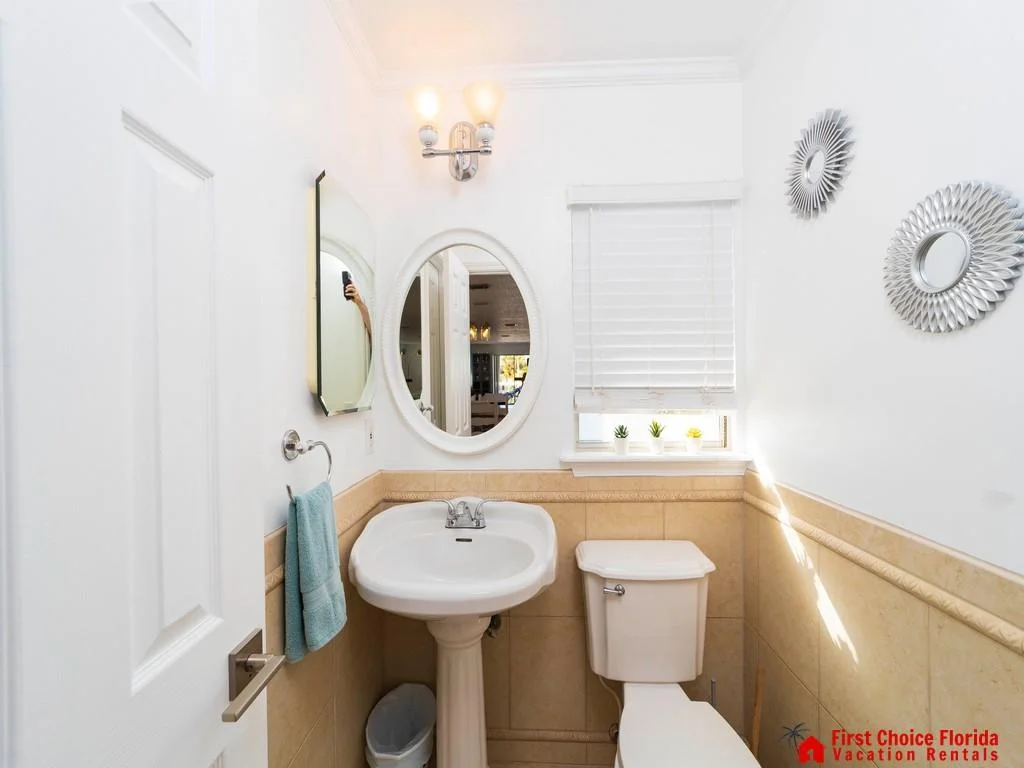 Half bath with beige tile wainscoting, pedestal sink, oval mirror, and small window with white blinds and plants.