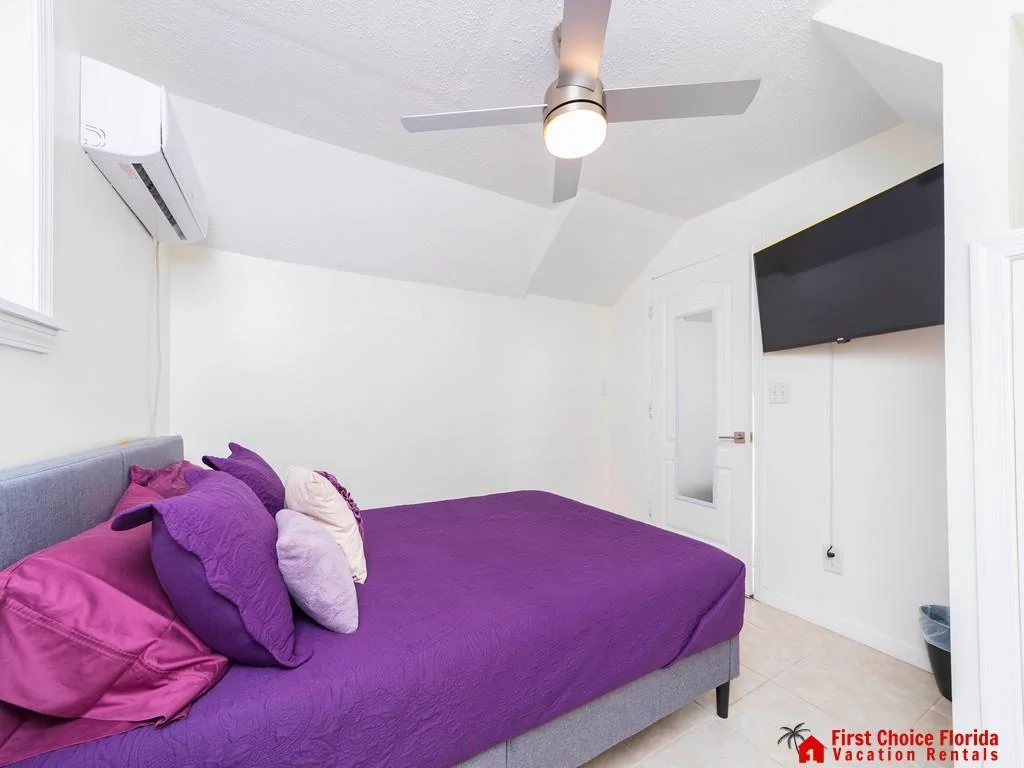Sloped ceiling with modern ceiling fan, white walls, and tiled floor complement the bed with layered purple pillows.