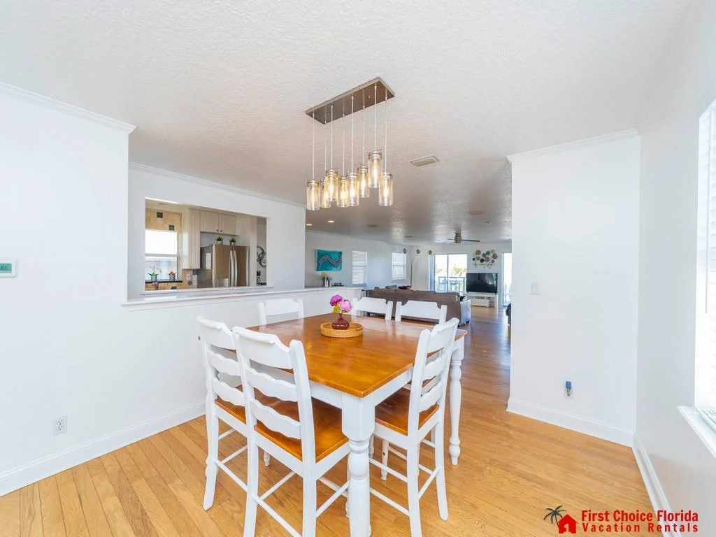 Dining area with farmhouse-style wooden table and white chairs, illuminated by a modern pendant light fixture.