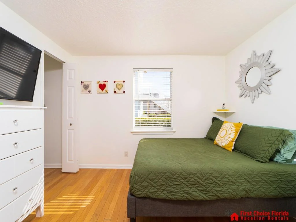 Natural light fills the bedroom with honey-toned hardwood floors, green quilted bed, and a white chest of drawers.