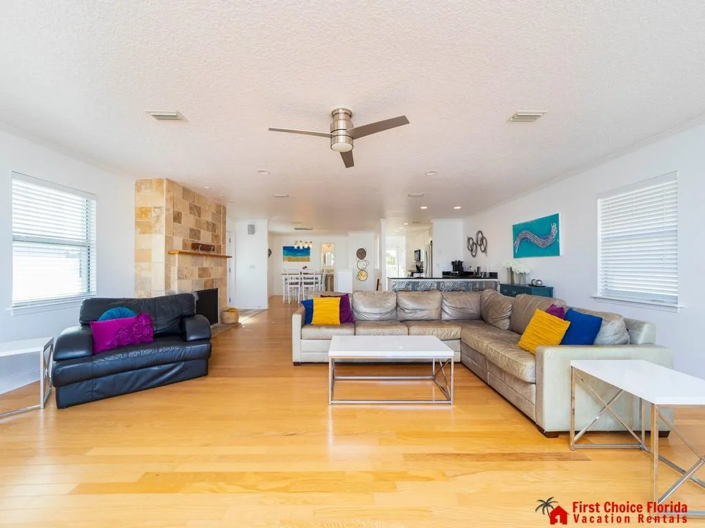 Spacious living room with honey-toned wood floors, leather sofas accented by colorful pillows, and a tiled fireplace.