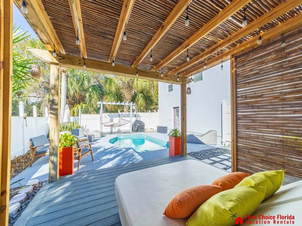 Shaded pergola with string lights overlooks a kidney-shaped pool, surrounded by gray decking and tropical plants.