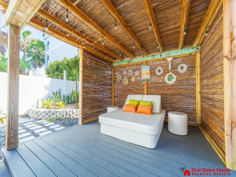 Covered outdoor lounge with bamboo slat walls, gray composite decking, string lights, and cushioned daybed with colorful pillows.