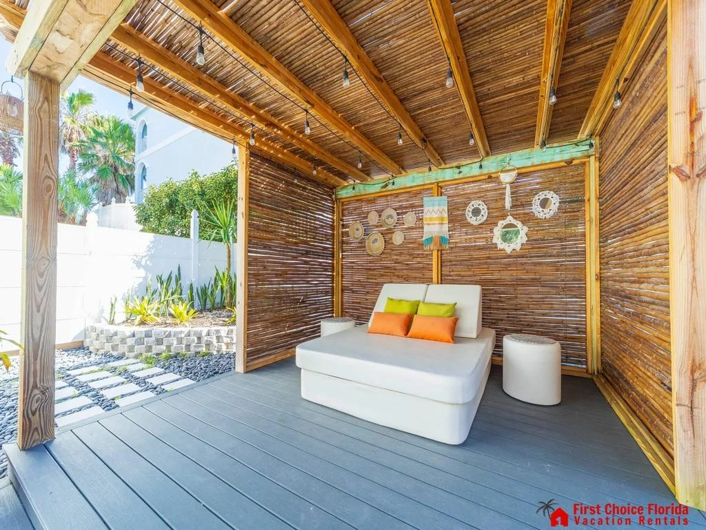 Covered outdoor lounge with bamboo slat walls, gray composite decking, string lights, and cushioned daybed with colorful pillows.
