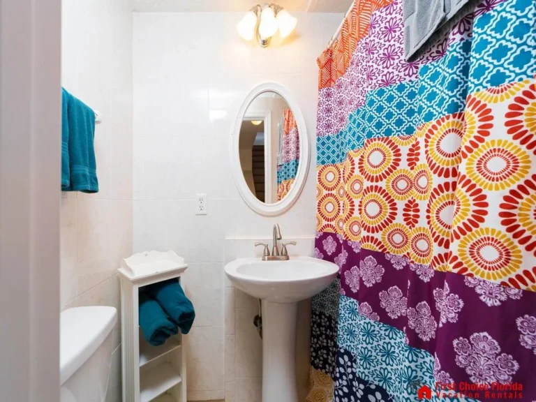 Small bathroom with white tile walls, oval mirror, pedestal sink, and colorful patterned shower curtain.