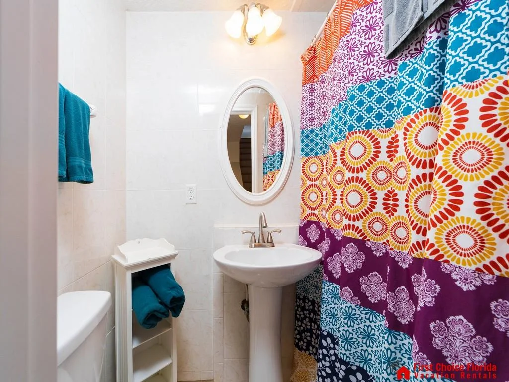 Small bathroom with white tile walls, oval mirror, pedestal sink, and colorful patterned shower curtain.