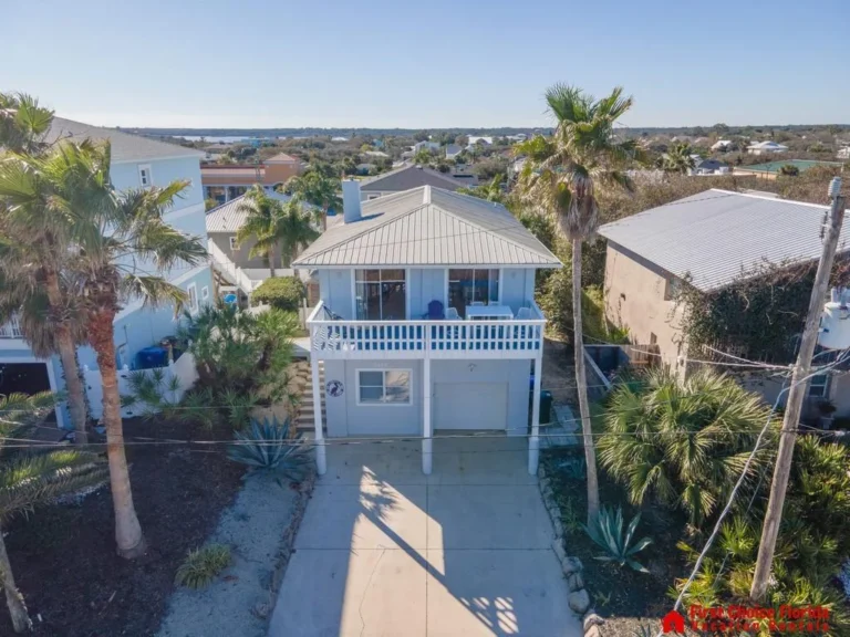 Two-story coastal home with white balcony railing, metal roof, concrete driveway, and palm trees in front yard.