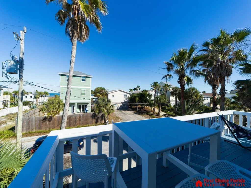Second-story balcony with white wooden railing, blue table, woven chairs, and partial ocean views beyond palm trees.
