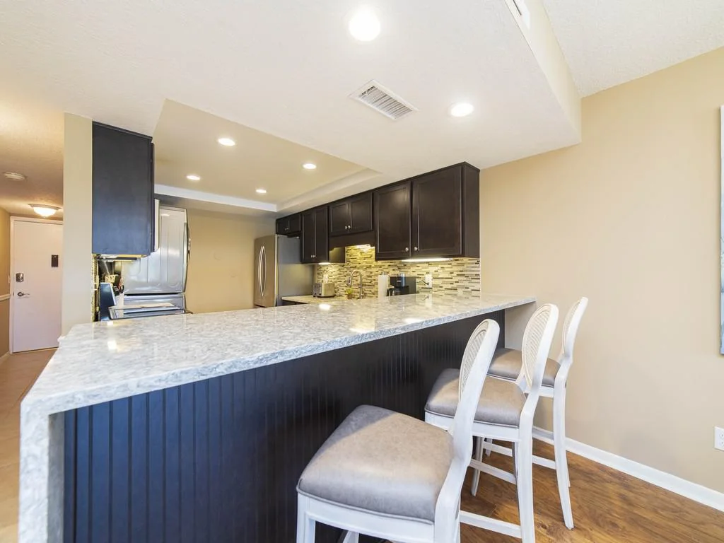 Marble countertop breakfast bar with cushioned white chairs faces dark wood cabinets and mosaic tile backsplash.