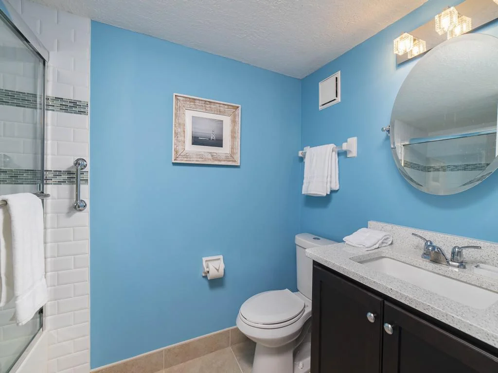 Bathroom with blue walls, white subway tile shower, dark wood vanity, round mirror, and overhead lighting.