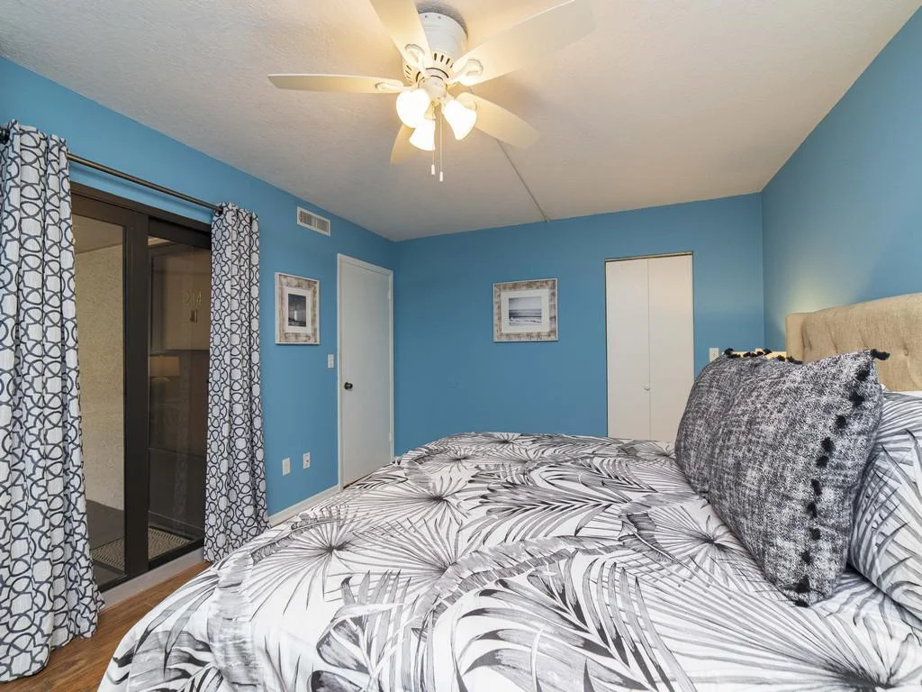 Blue-walled bedroom with patterned curtains, white ceiling fan, and a bed dressed in black-and-white botanical linens.