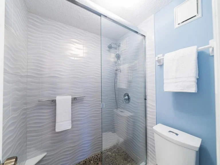 Shower area with textured wave-patterned white tiles, pebble stone floor, glass sliding door, and blue painted wall.