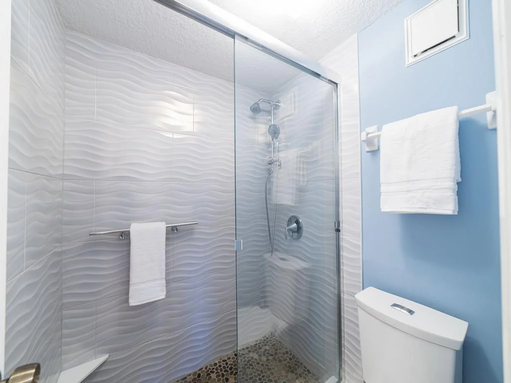 Shower area with textured wave-patterned white tiles, pebble stone floor, glass sliding door, and blue painted wall.