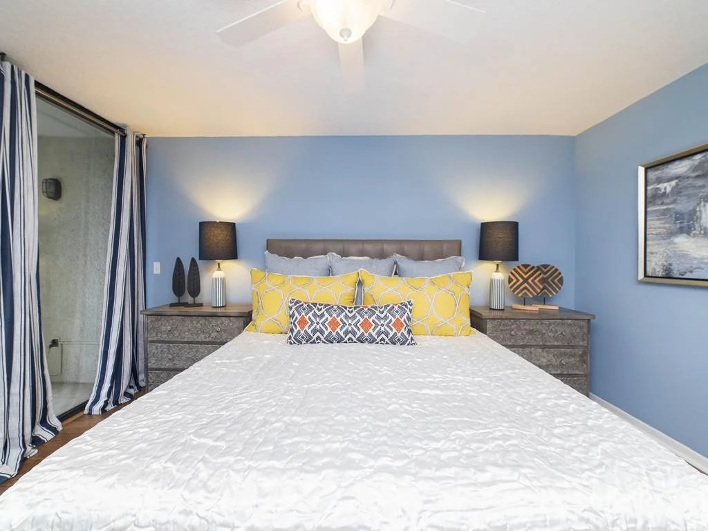 Blue walls frame the bedroom’s queen bed with yellow and patterned pillows, flanked by dark wood nightstands and lamps.
