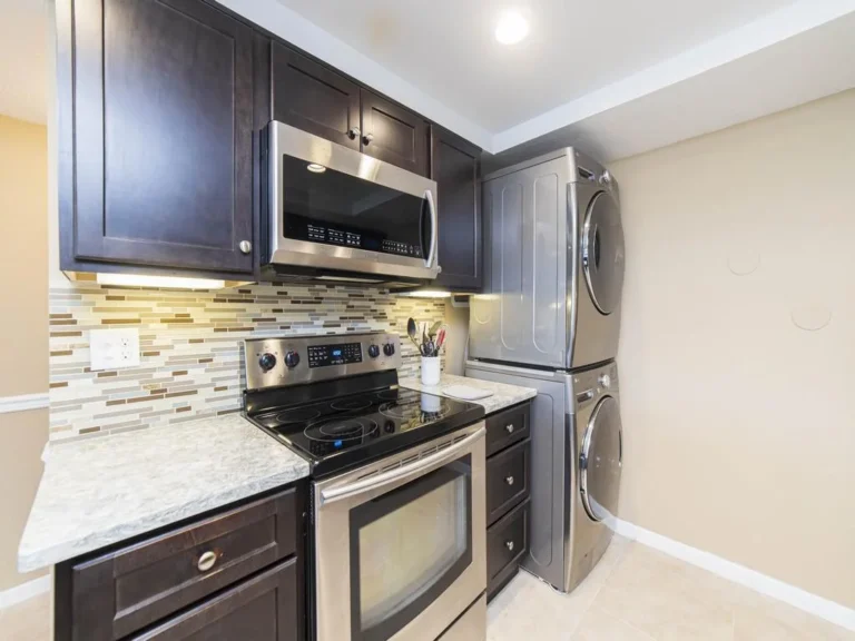 Stacked stainless steel washer and dryer sit next to a black electric stove and dark wood cabinets with a tile backsplash.