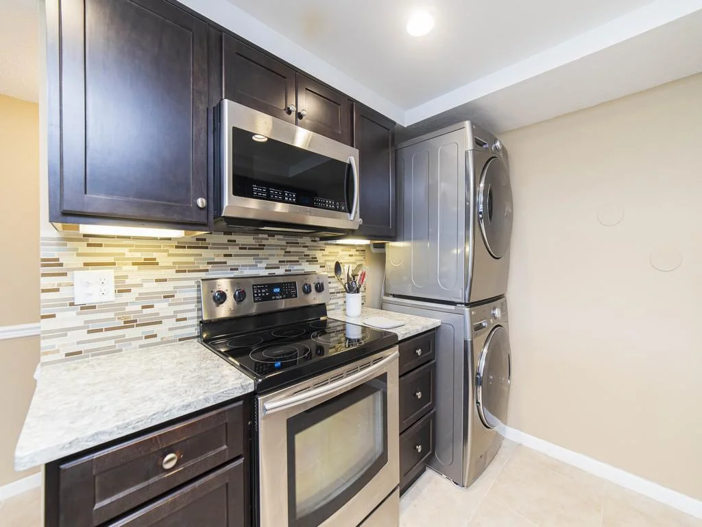Stacked stainless steel washer and dryer sit next to a black electric stove and dark wood cabinets with a tile backsplash.