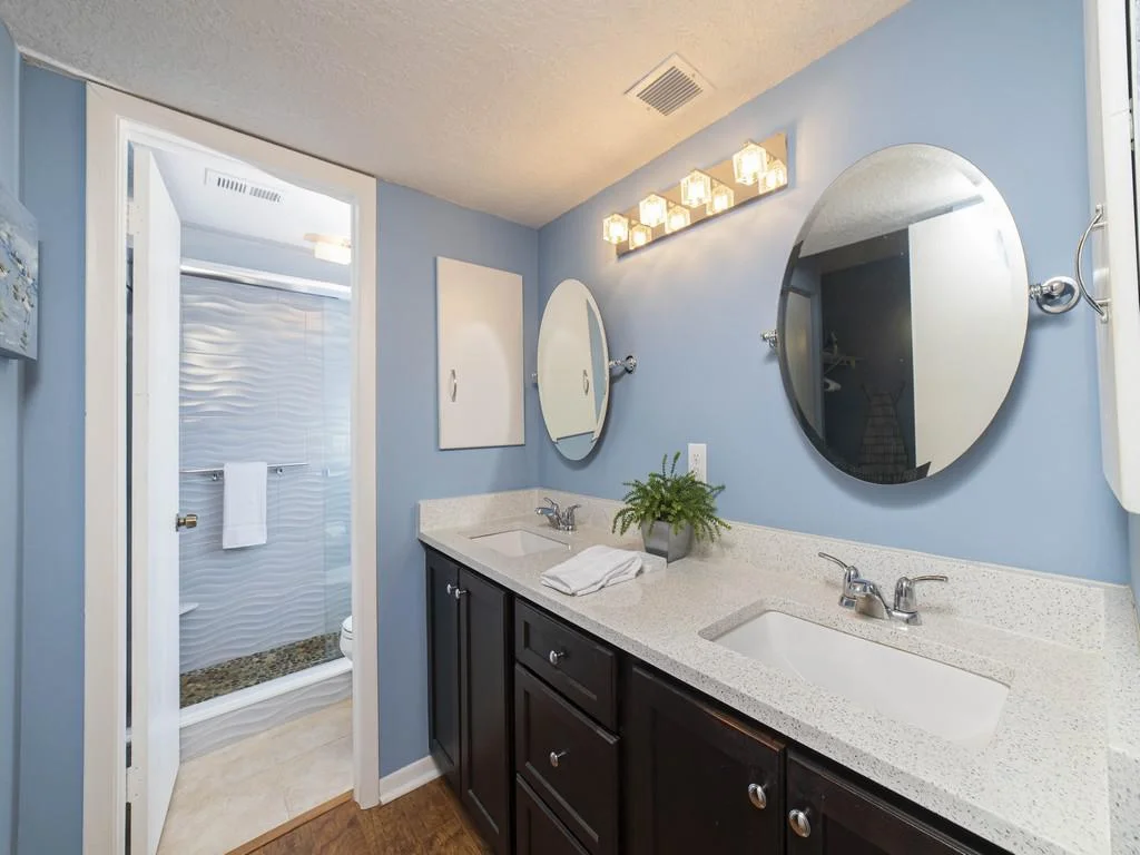 Double vanity with oval mirrors and quartz countertops in a blue-painted bathroom with a walk-in shower.