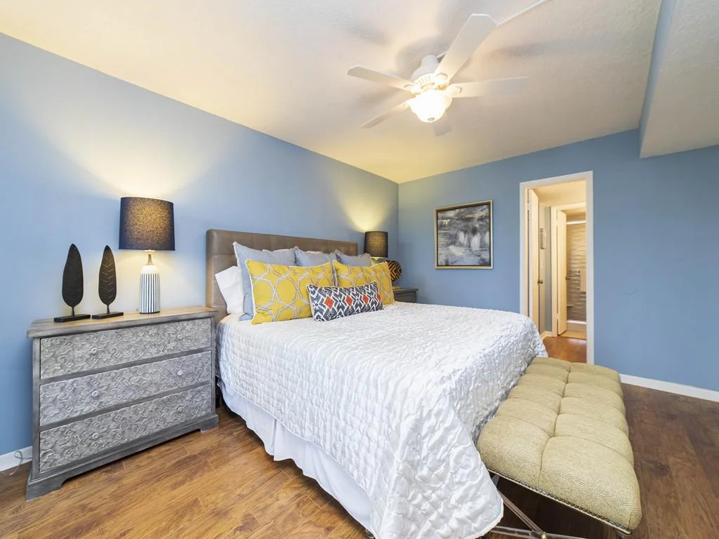 King bed with white quilt and yellow pillows sits against blue walls beside a textured gray dresser and bench.