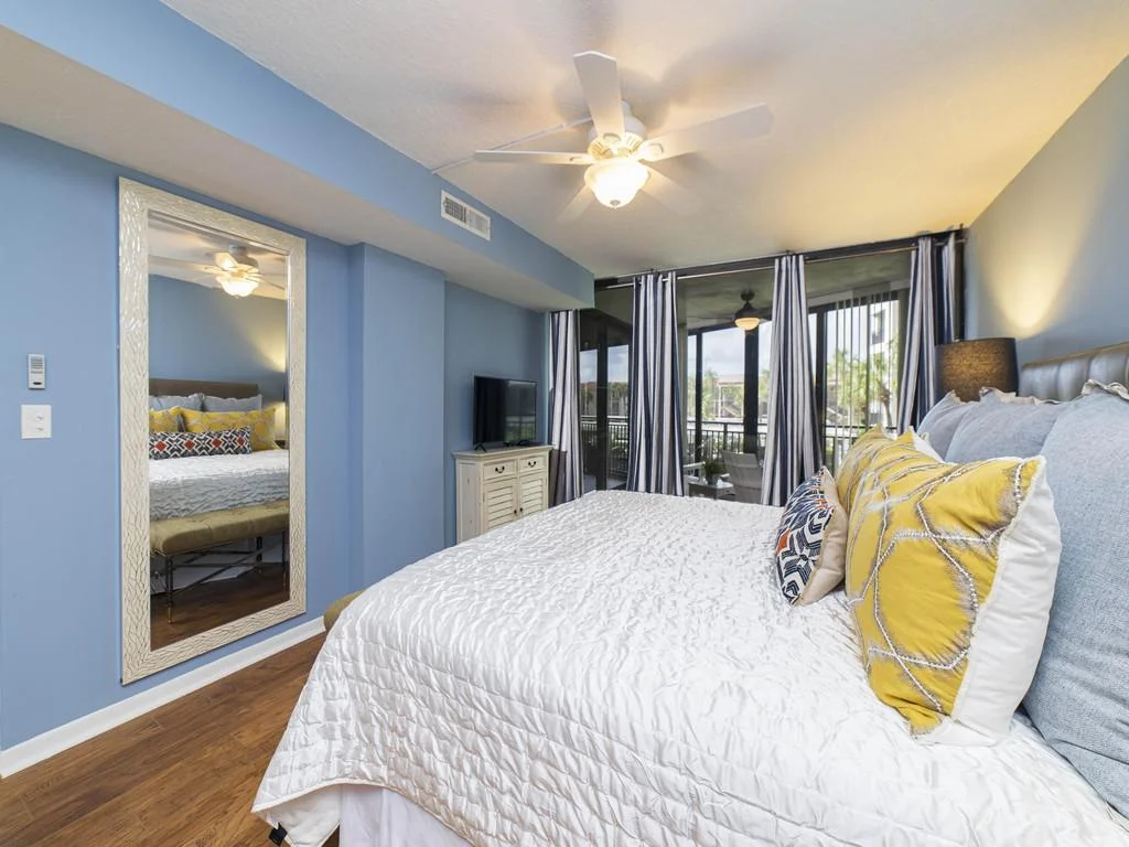 Blue walls frame a king bed with yellow and patterned pillows, a ceiling fan, and sliding glass doors to the balcony.