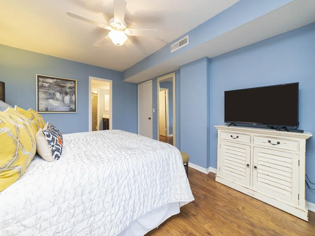 Blue-walled bedroom with white quilted bedding, yellow and patterned pillows, ceiling fan, and TV on distressed cabinet.
