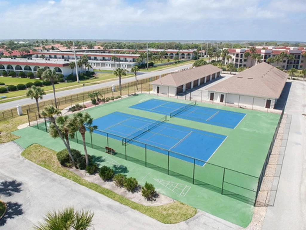 Two adjacent blue tennis courts with green surrounding surface, enclosed by chain-link fencing and nearby palm trees.